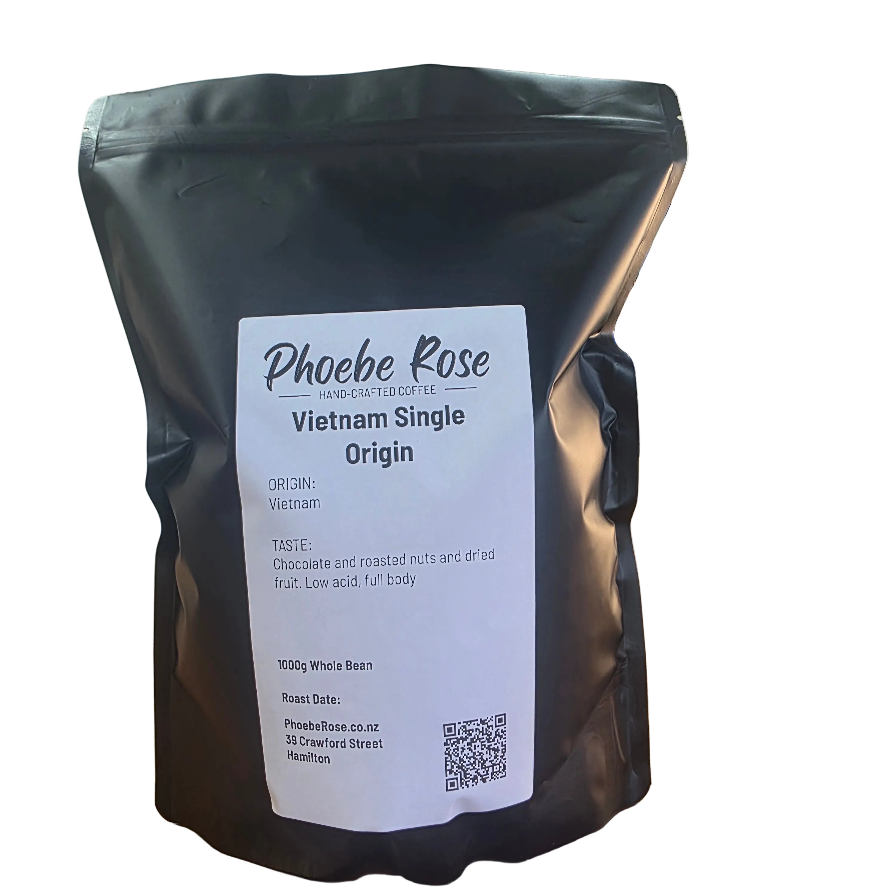 A black stand-up coffee bag labeled 'Phoebe Rose Vietnam Single Origin' standing on a wooden table in a home interior with a lamp, chair, and plant visible in the background.